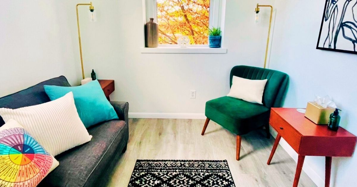 Cozy living room: grey sofa with colorful pillows, emerald green chair, red side table, window with fall foliage.
