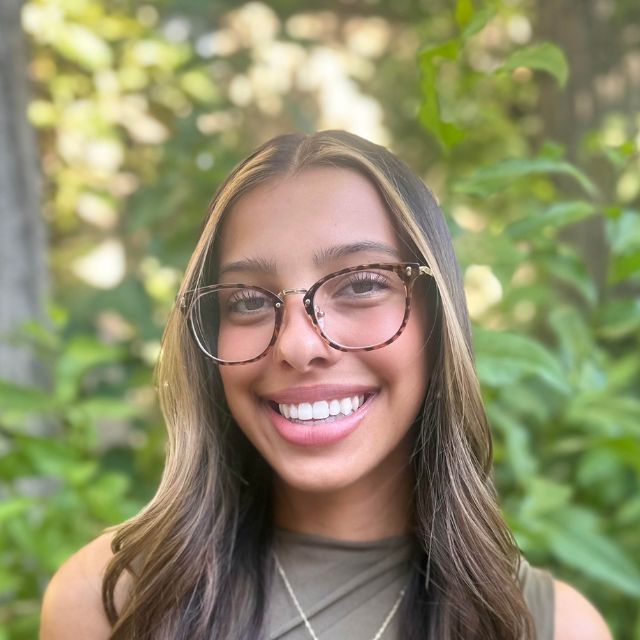 Young woman with glasses smiles at the camera, hair highlights, outdoor green backdrop.