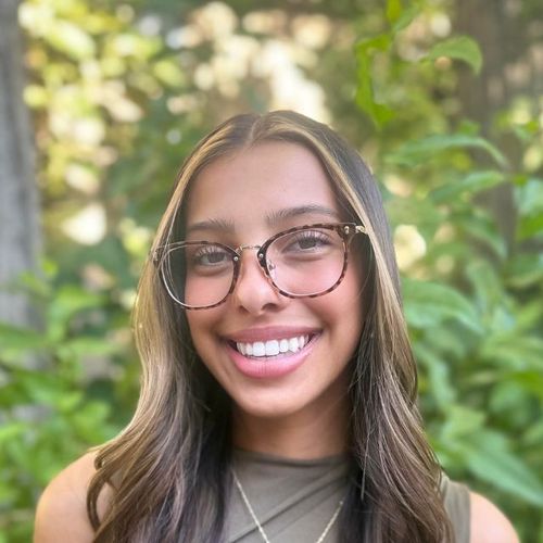 Young woman with glasses smiles at the camera, hair highlights, outdoor green backdrop.
