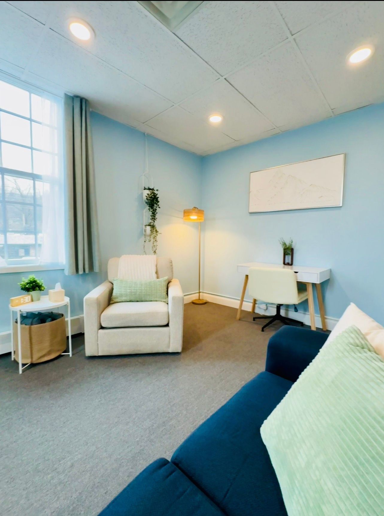 Light blue therapy office with a couch, armchair, desk, and window.