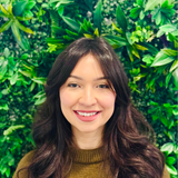 Woman smiling, long dark hair, wearing a brown sweater, in front of a green leafy wall.
