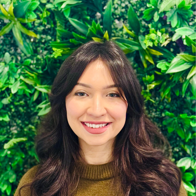 Woman smiling, long dark hair, wearing a brown sweater, in front of a green leafy wall.