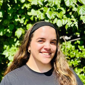 Woman with long brown hair wearing a black headband smiles in front of green foliage.