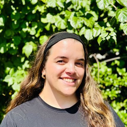 Woman with long brown hair and a black headband smiling in front of green foliage.
