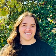 Smiling person with long wavy brown hair wearing a black sweater, standing in front of leafy green trees.