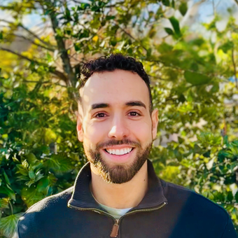 Man with a beard smiling in front of green foliage, wearing a black jacket.
