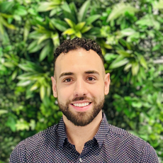 Woman with curly brown hair, smiling, wearing a pink jacket, in front of green foliage.