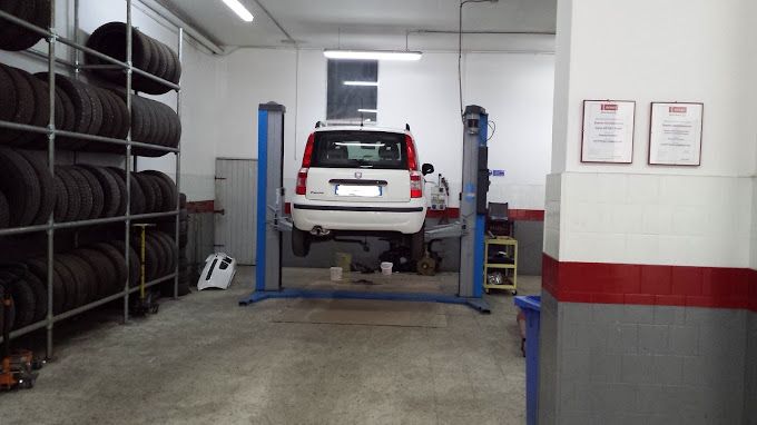 Un SUV bianco su un ponte sollevatore in un'officina meccanica, con pneumatici impilati lungo la parete sinistra.