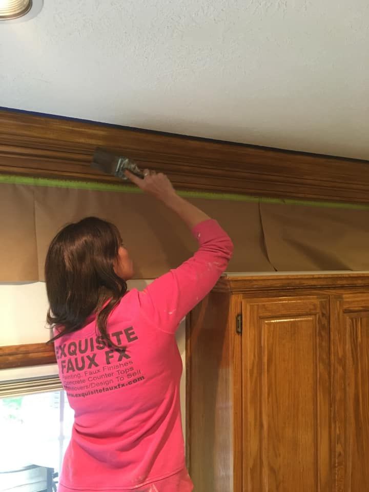 Woman painting wooden trim, standing on a step stool, in a kitchen. The trim is taped off, and she is wearing pink.