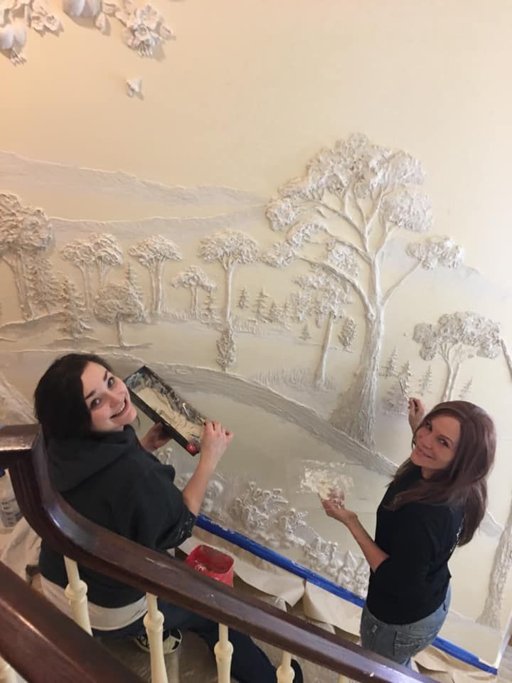 Two people smile while applying a white sculptural plaster relief of a forest landscape onto a wall beside a staircase.