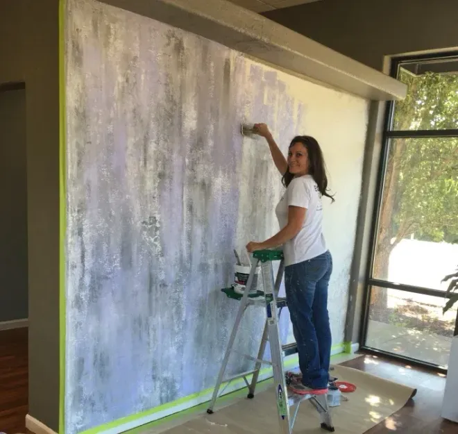 A person on a ladder paints a textured, gray and purple focal wall in a room with a large window.