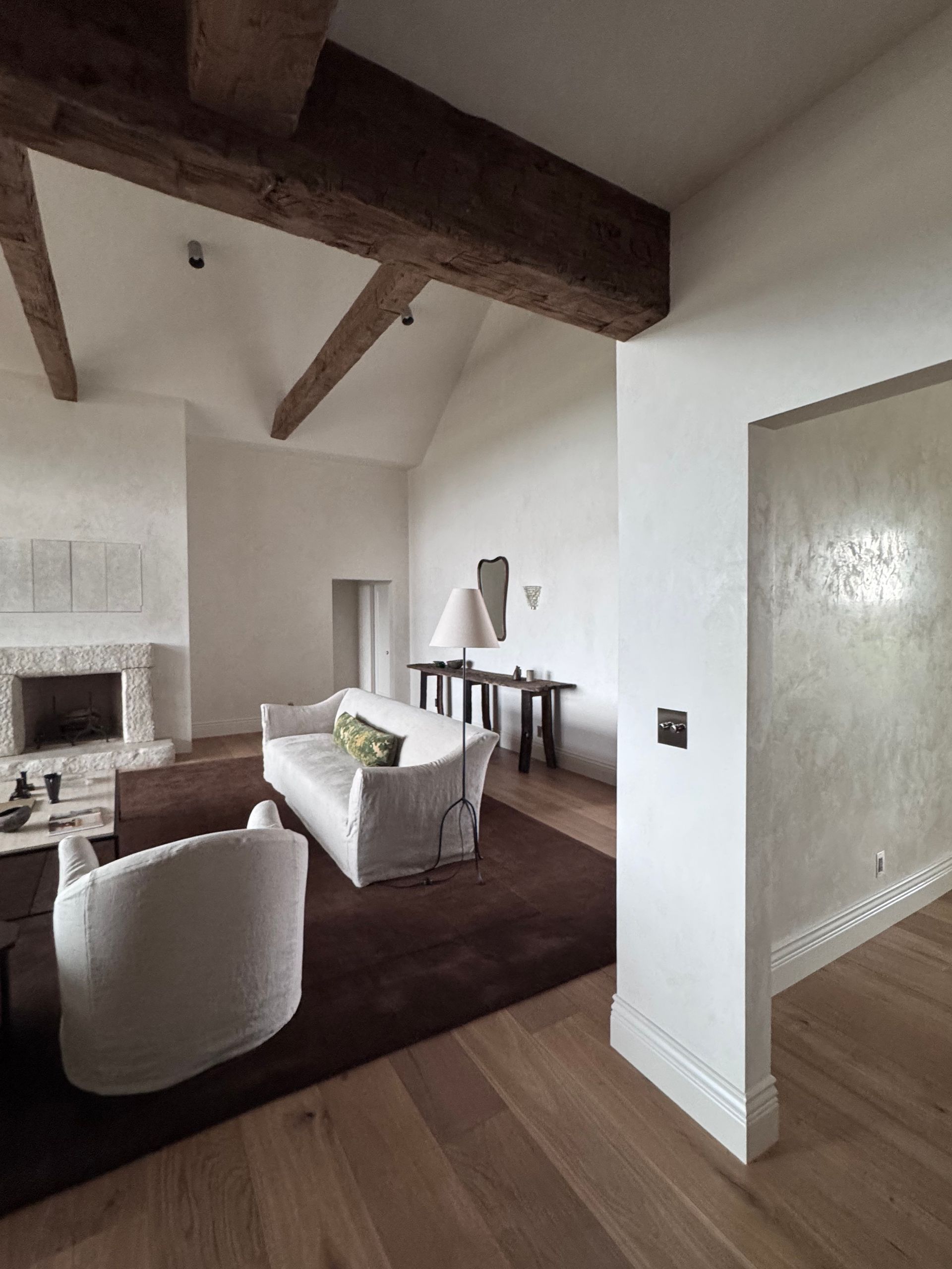Living room with exposed wood beams, white walls, and a stone fireplace. Furnishings include white sofas and a dark brown rug.