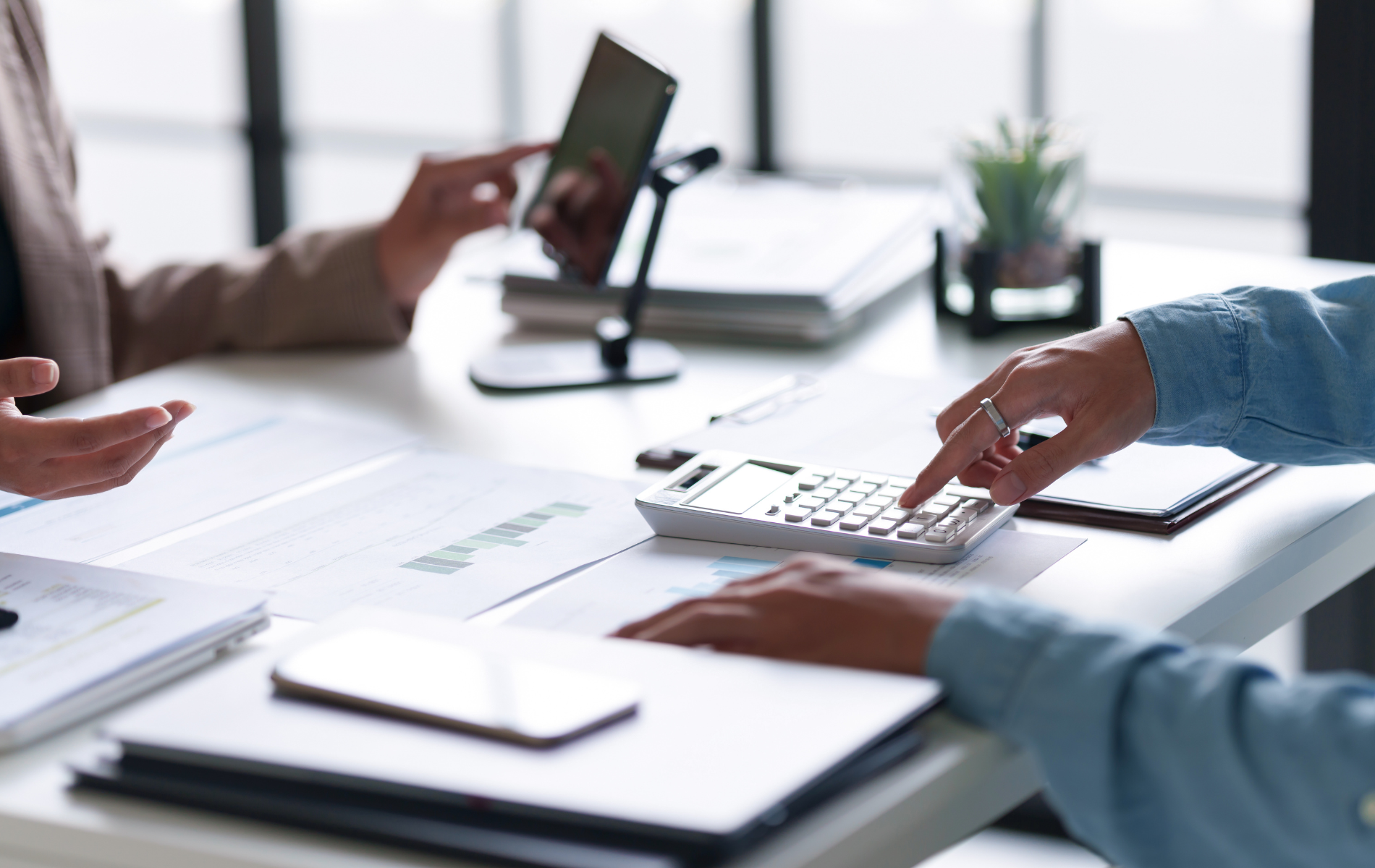 People calculating finances with documents, calculator, and tablet on a white table.