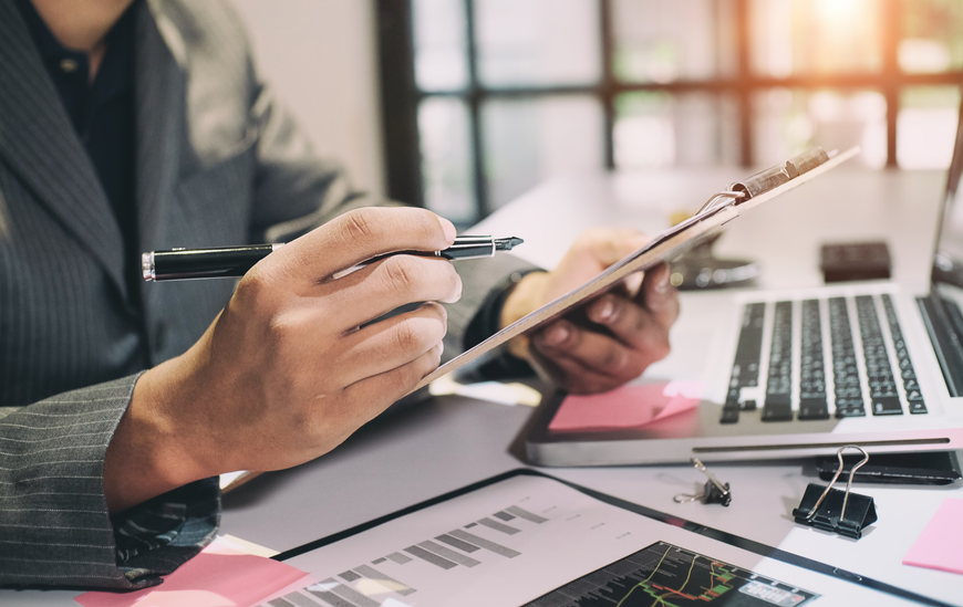 Person in a suit reviews documents on a clipboard with a pen, next to a laptop and financial charts.