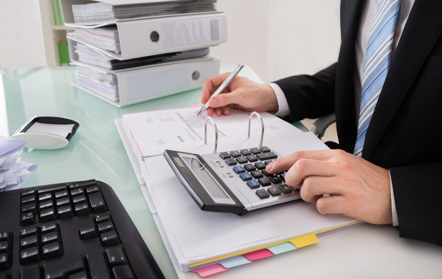Person in a suit calculating with a calculator, writing on papers at a desk with binders and a keyboard.