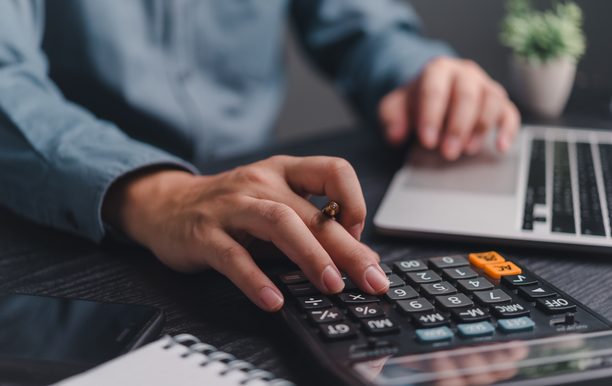 Hands using a calculator with a laptop and notebook on a desk.