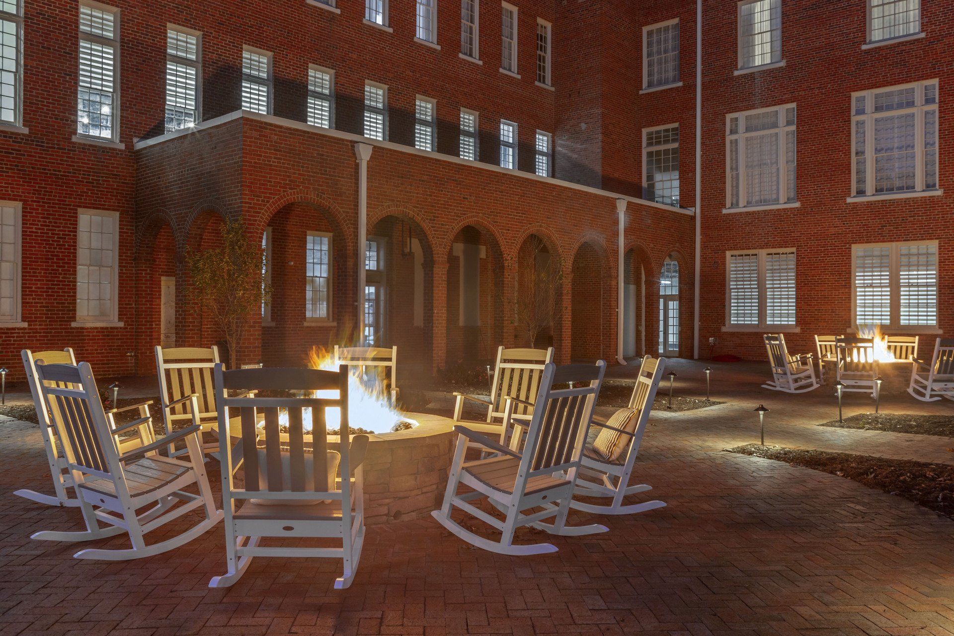 A group of rocking chairs are sitting around a fire pit in front of a brick building.
