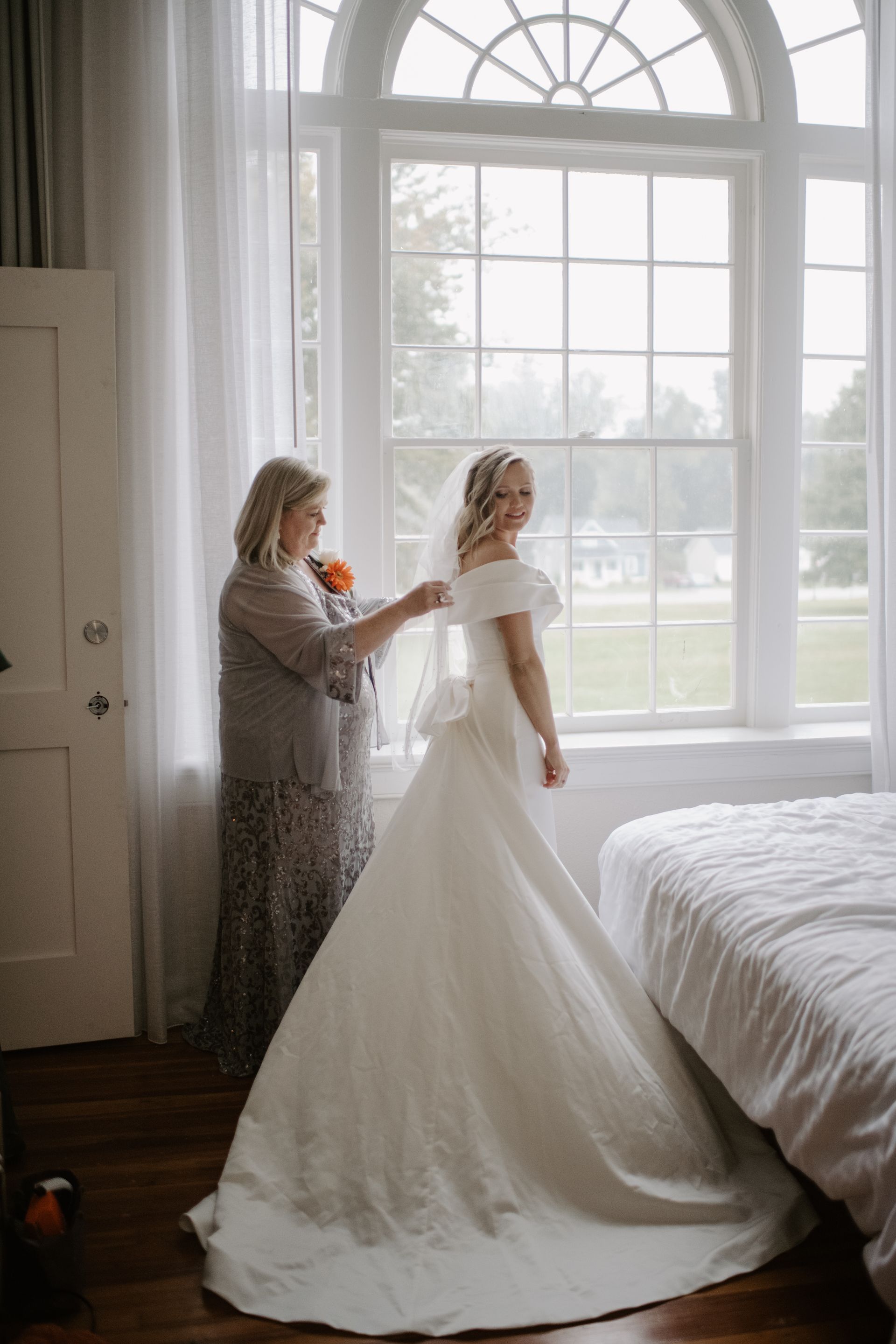 A woman is helping a bride get ready for her wedding in front of a window.