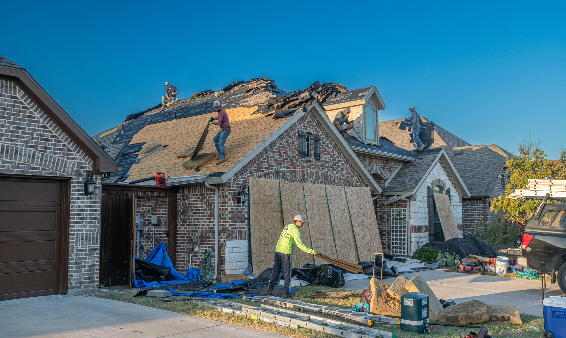 Roofers removing old shingles from a house; one on the roof, two on the ground.