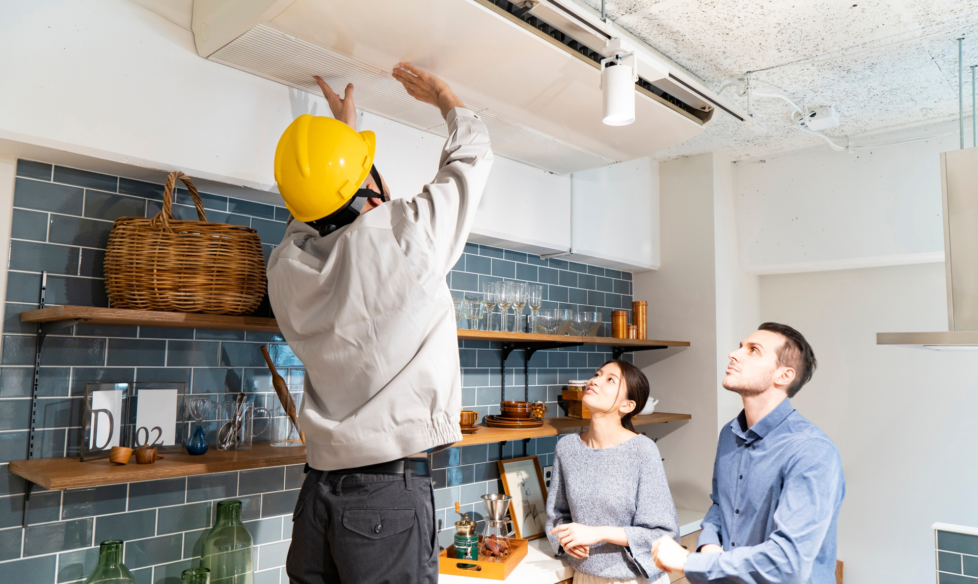 Man in hard hat installing ceiling fixture while couple watches in kitchen.