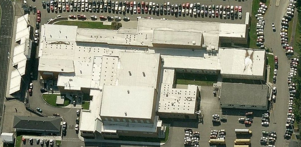 Aerial view of a large industrial building with multiple white roofs, surrounded by parking lots and trees.