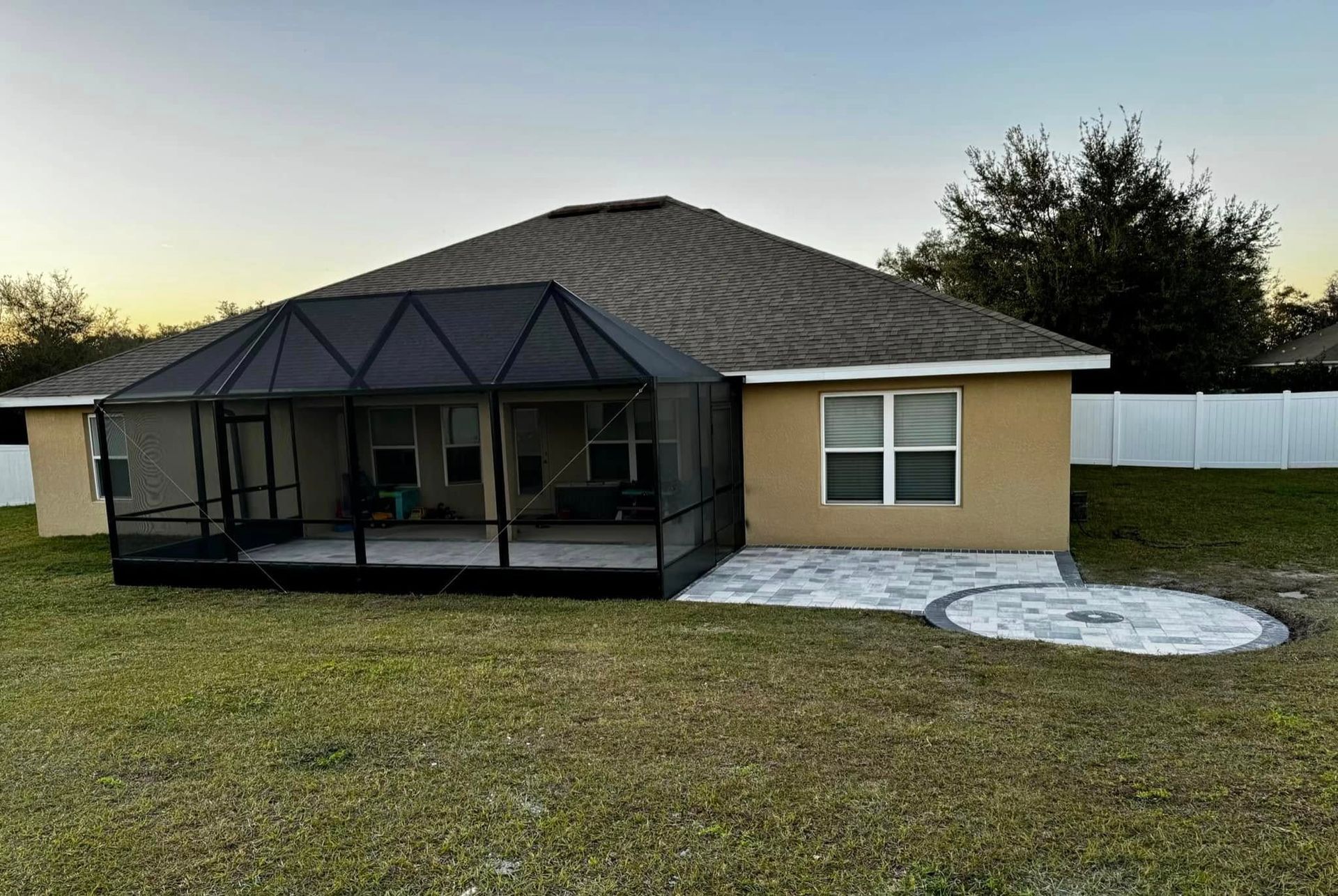 Backyard with a screened-in patio, beige house, and a circular stone feature. Green grass and white fence.