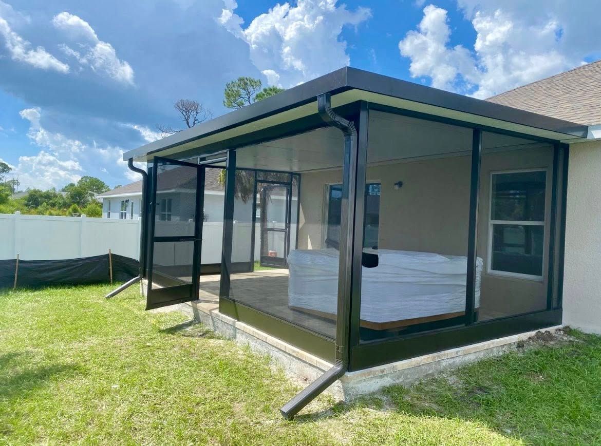 Screened patio extension with black frame and gutter, attached to a beige house, green grass, blue sky.