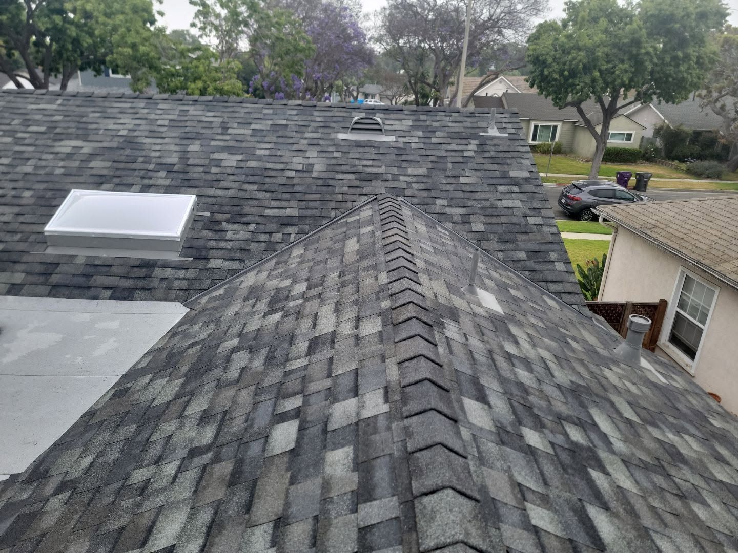 Roof shingles on a house, viewed from the ridge with a skylight and neighboring homes in the background