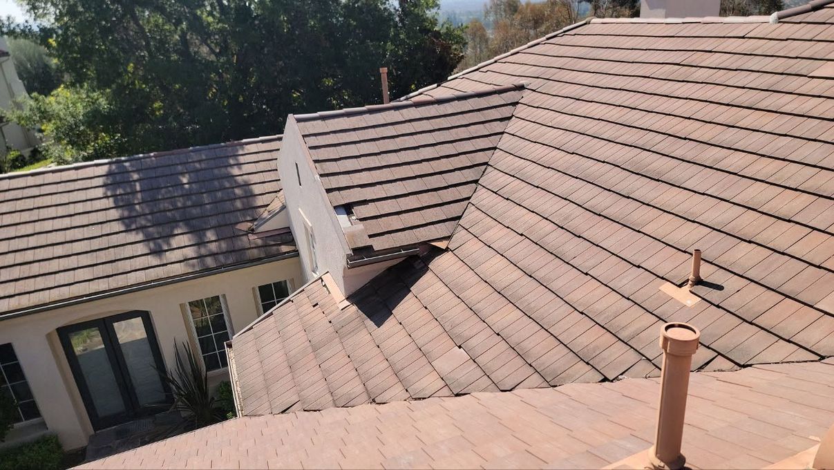 Sunlit residential rooftops with tan shingles and a white dormer, viewed from above