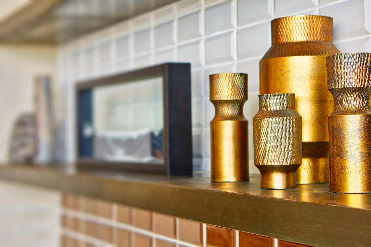Gold decorative vases on a shelf with a framed picture against a tiled wall.