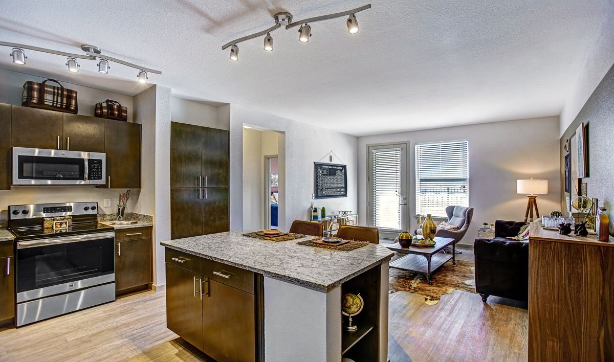 Modern kitchen and living area with a granite island, stainless steel appliances, and wood floors.