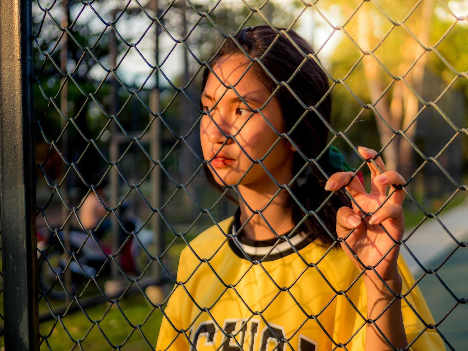 An Asian teenage girl standing behind a fence looking pensive.