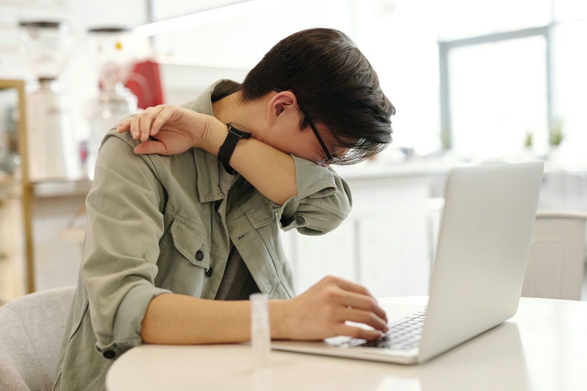 Person at a white desk using a laptop, leaning on one arm and rubbing their shoulder.