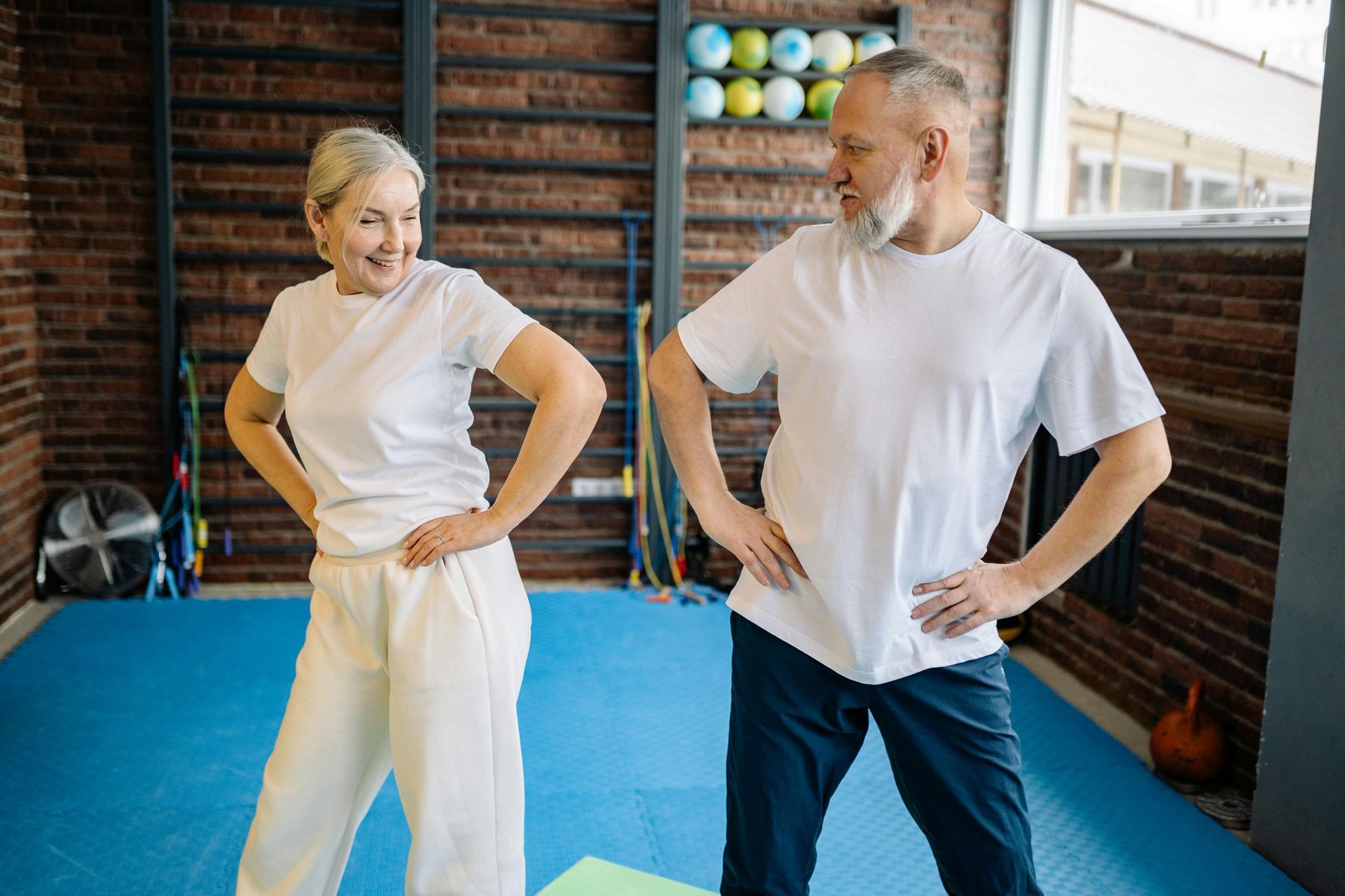 Two people in white shirts and workout pants standing hands on hips in a gym studio