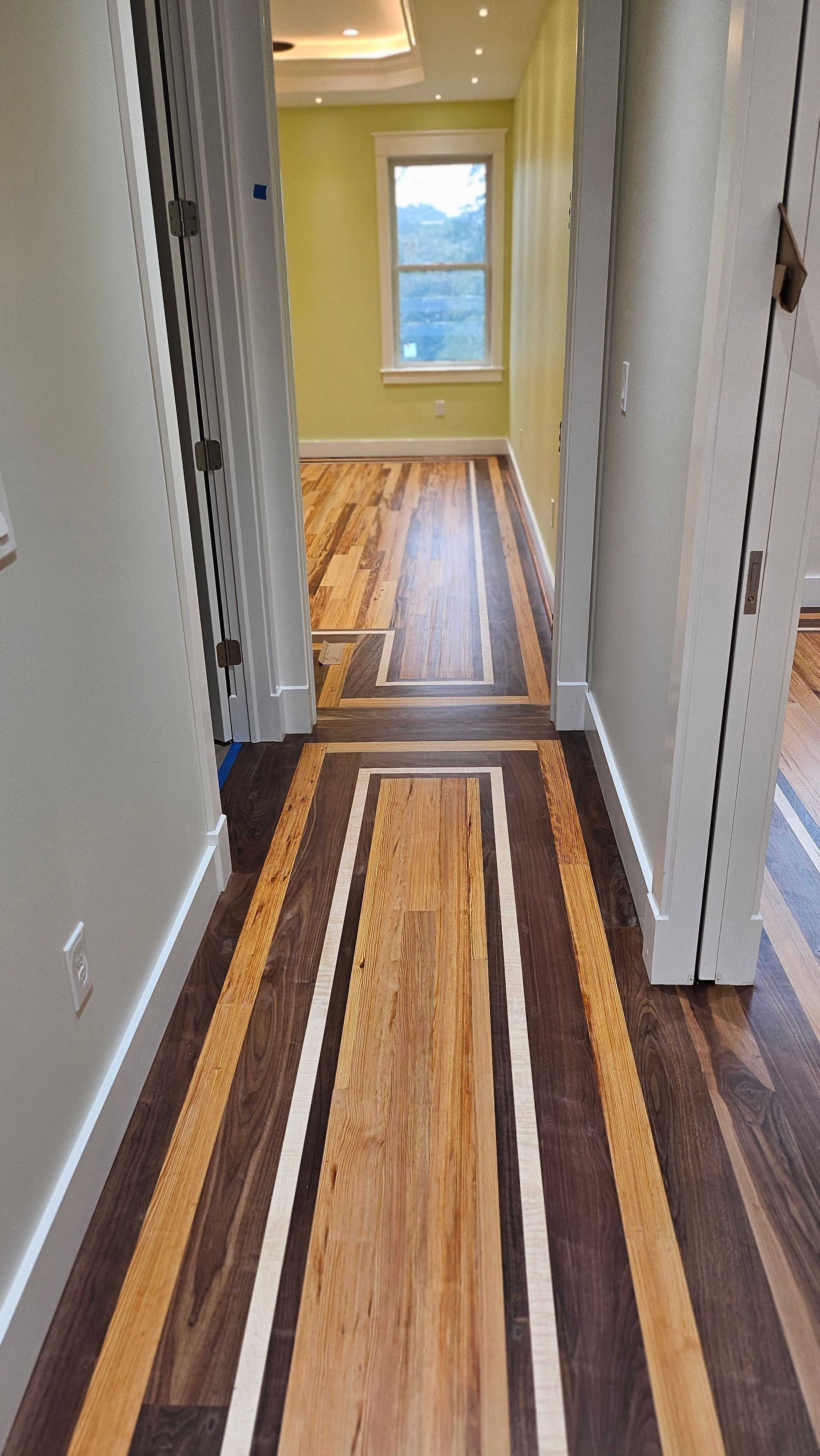 Hallway with a hardwood floor featuring a central light wood design framed by darker wood strips and white borders. Yellow walls at the end of the hall.