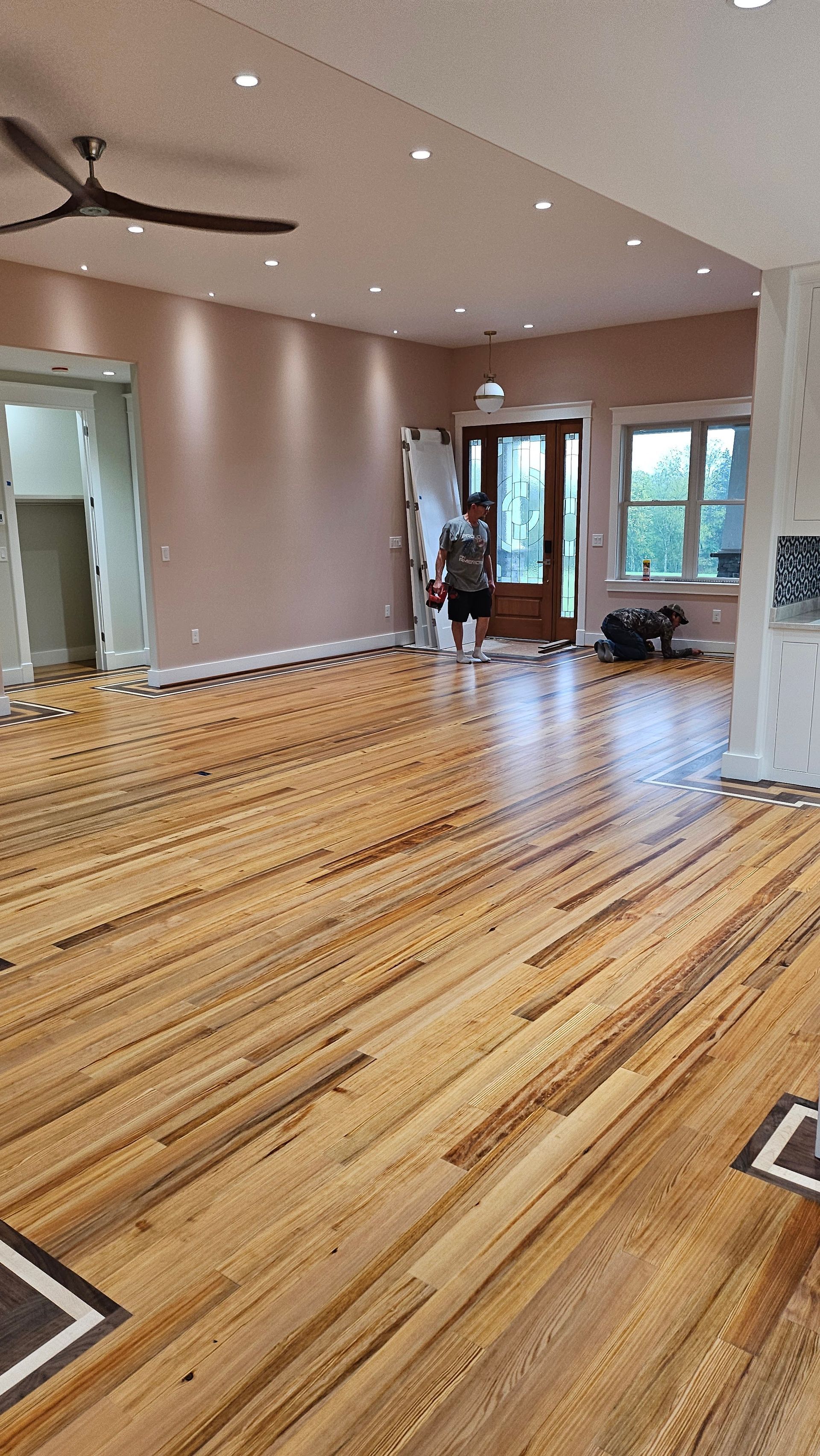 Wide shot of a spacious room with hardwood floors, light pink walls, and a dark ceiling fan. A doorway leads to another room on the left.