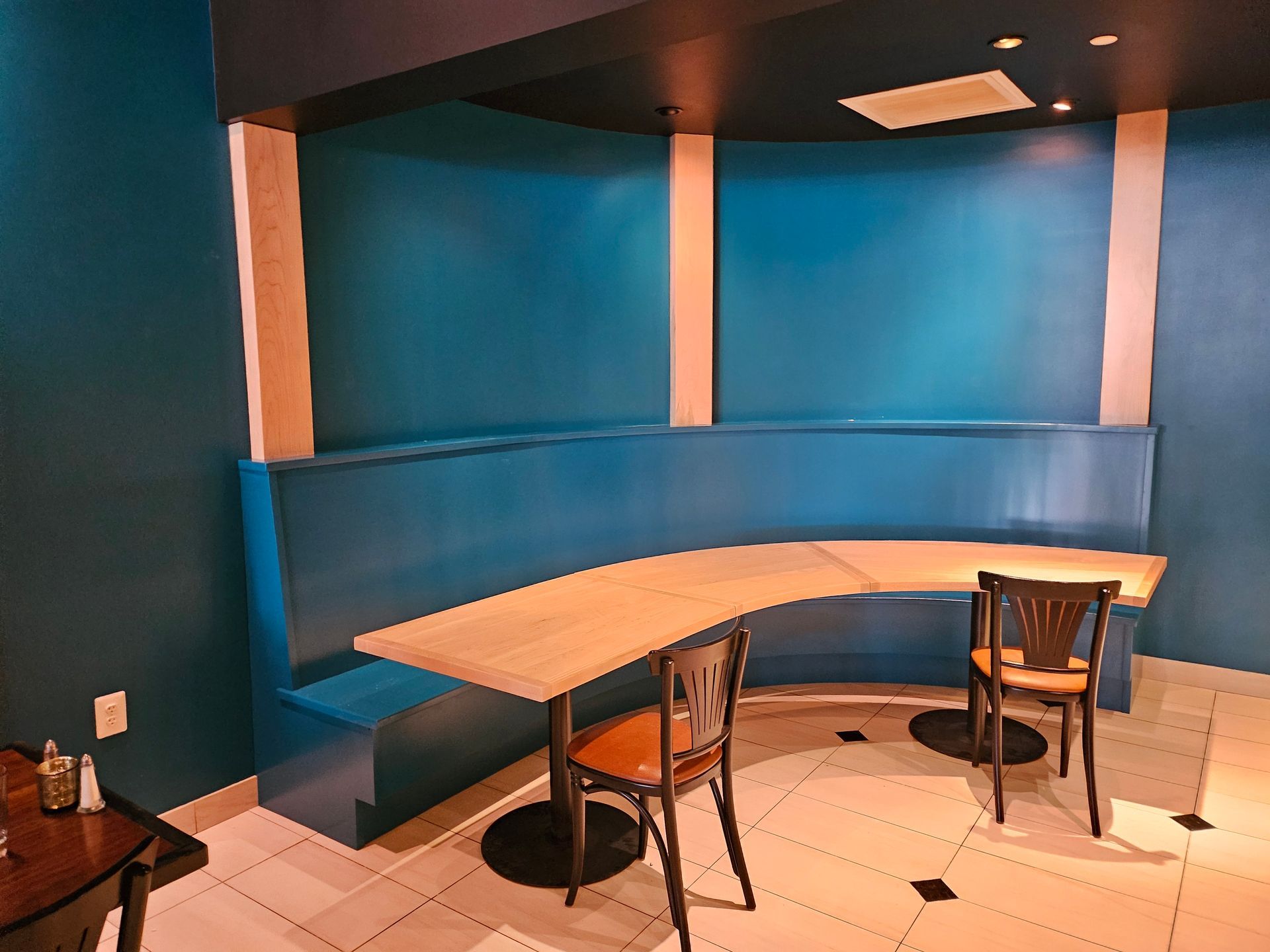 Booth seating in a restaurant with a teal blue wall and light wood table. Two chairs are positioned at the table.