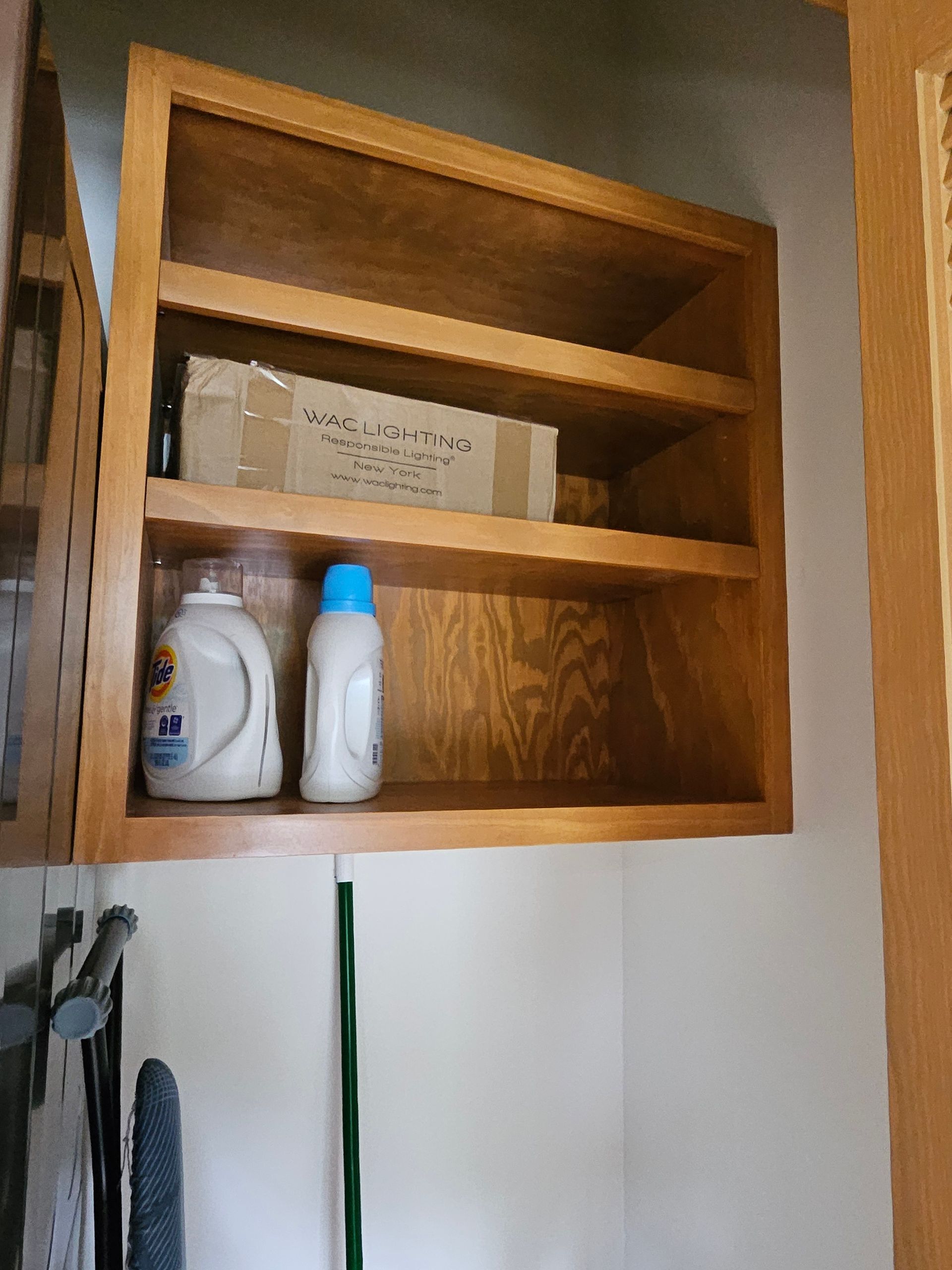 Wooden cabinet with two shelves holding detergent bottles and a cardboard box, mounted on a white wall.