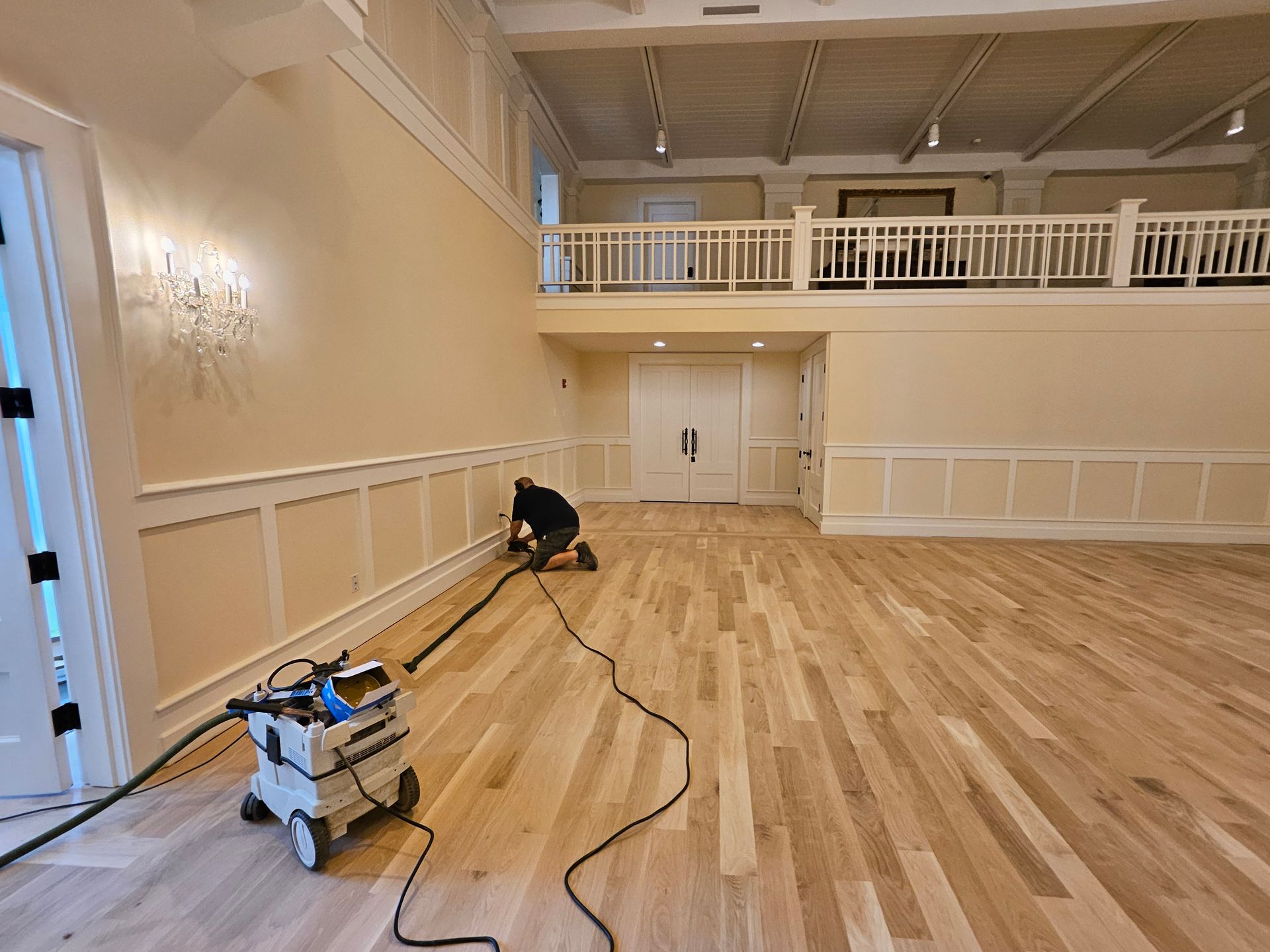 A person using a machine to sand hardwood floors in a large, empty room with white walls and wainscoting.
