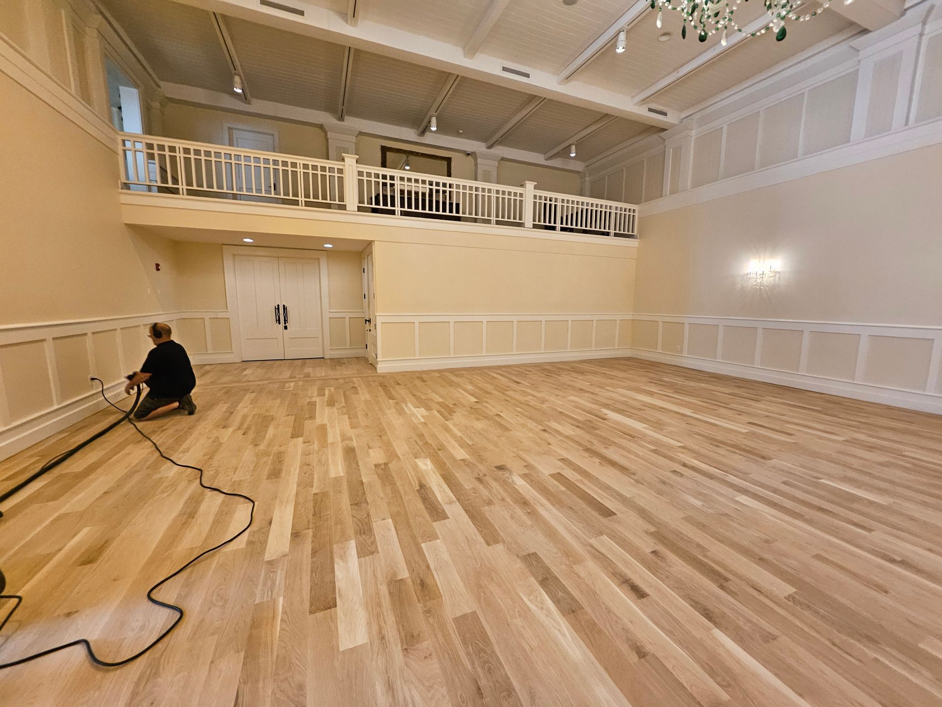 A person works on a light wood floor in a large, empty room with a balcony and wainscoting.
