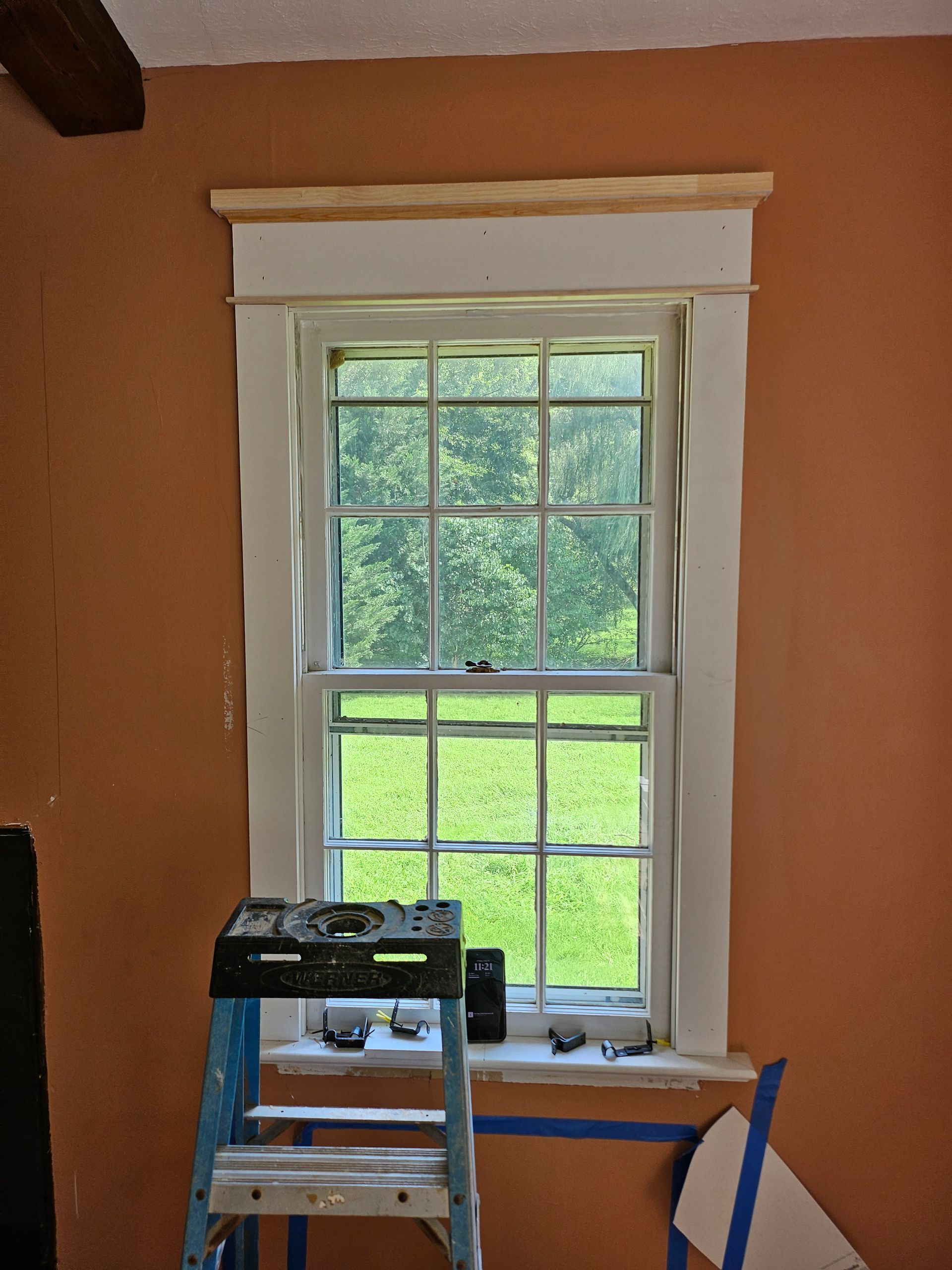 A tall window framed in white against an orange wall. A wooden shelf with trim sits above the window. A ladder is in front of the window.