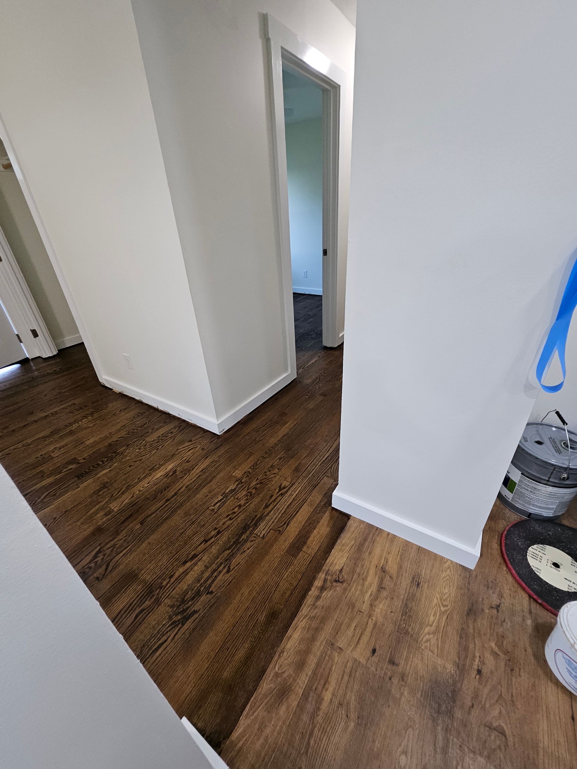 Brown wooden floor in a hallway with white walls, a doorway, and cleaning supplies.
