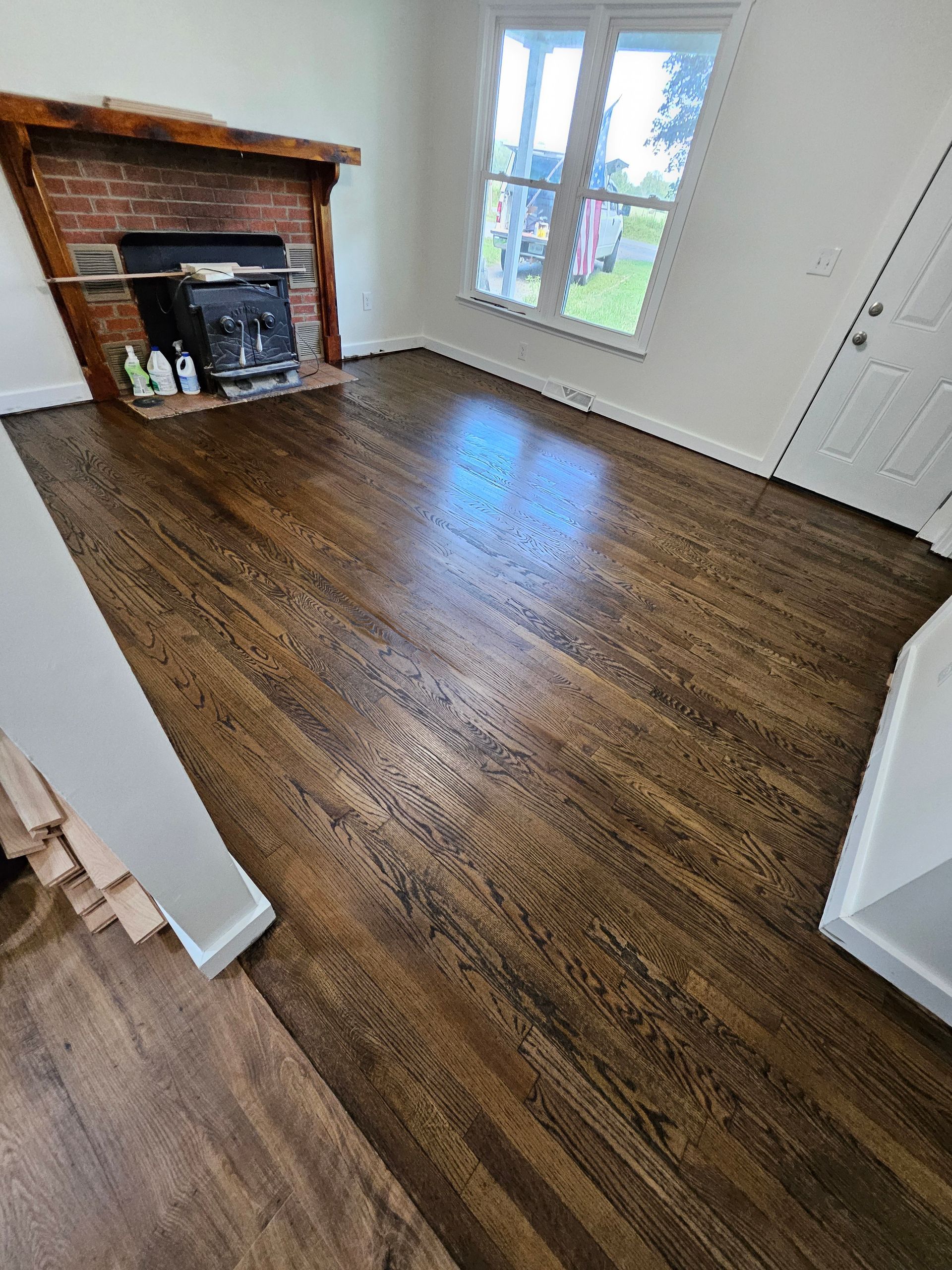 Newly finished hardwood floors in a living room with a brick fireplace and white walls, a window, and a door.