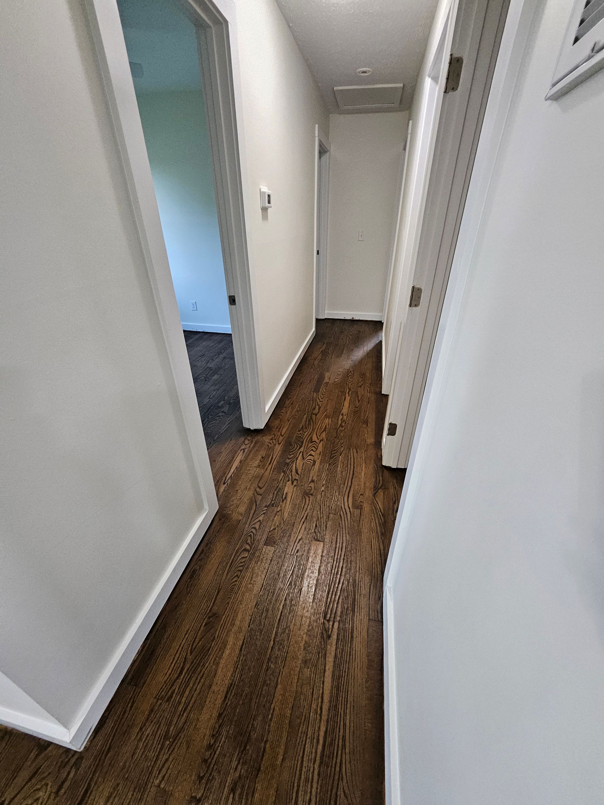 Narrow hallway with dark wood flooring, white walls, and closed doors. One open door leads to a room with carpet.