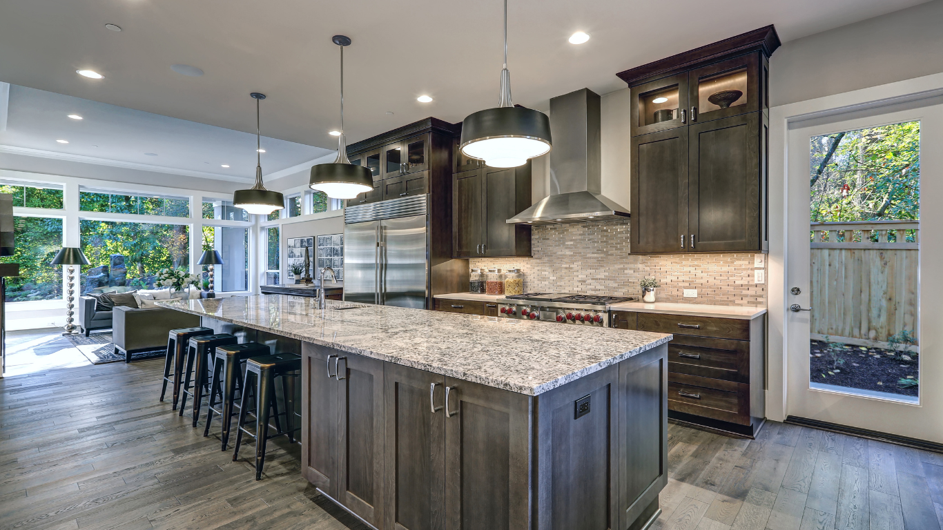 Modern kitchen with a large island, dark cabinets, and a stainless steel range hood. Natural light floods the space.