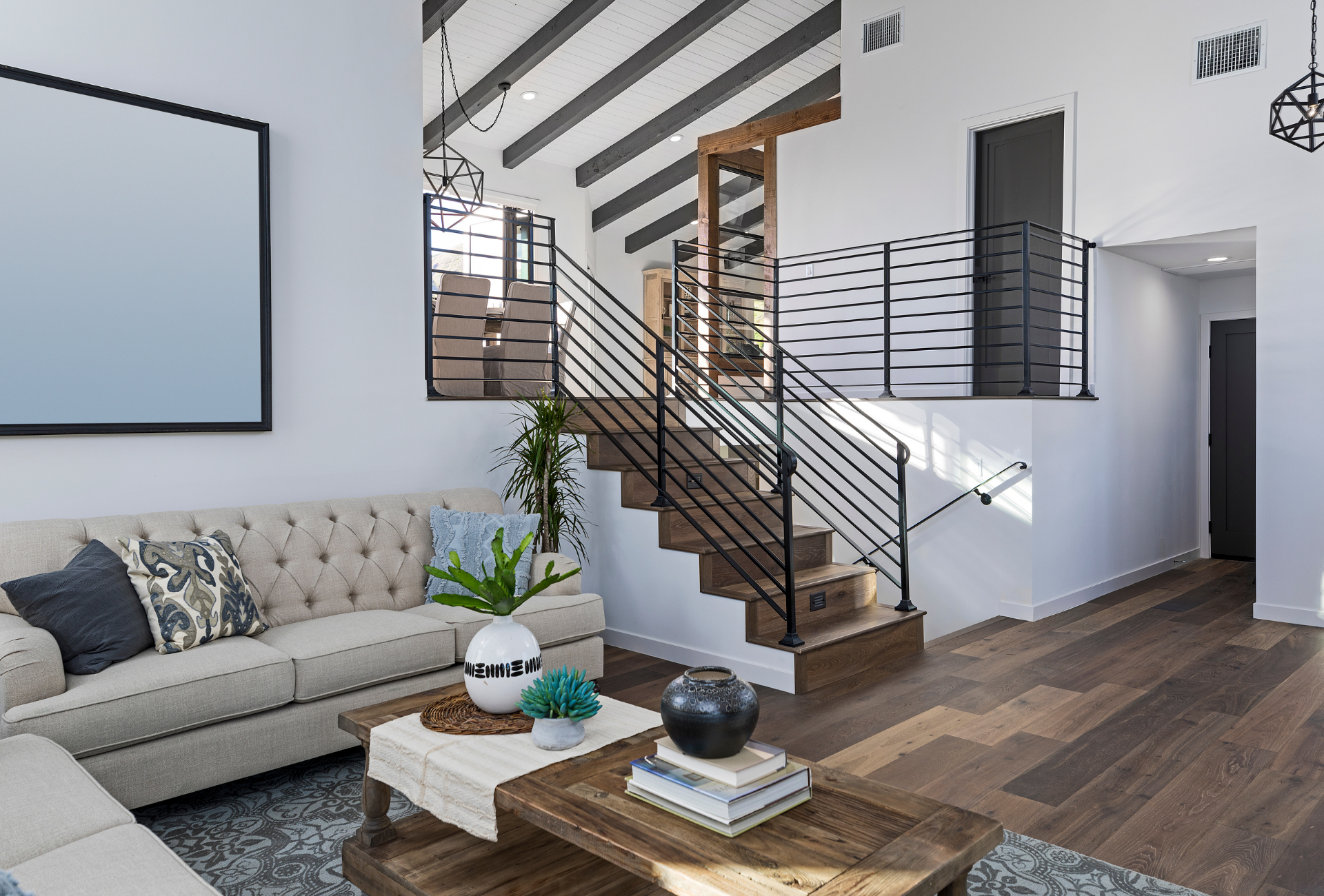 Living room with staircase, tufted beige sofa, wooden coffee table, and hardwood floors. White walls and black railing.