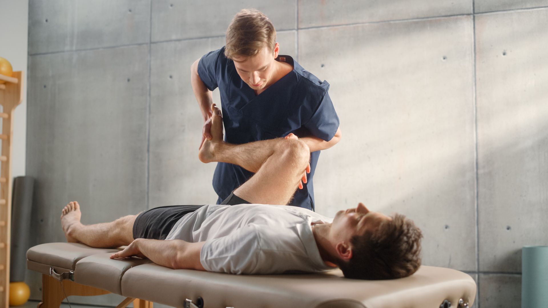 A therapist guides an athlete’s leg through a stretching exercise on a treatment table.