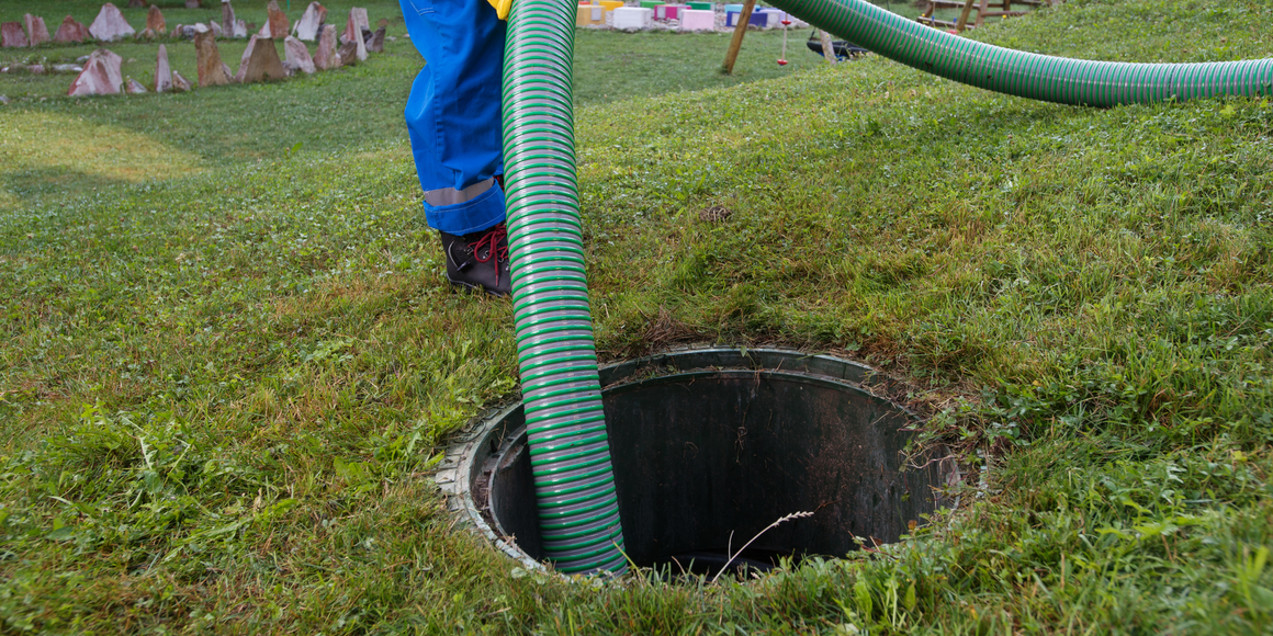 A person in blue overalls with a green hose servicing a septic tank in a grassy area.