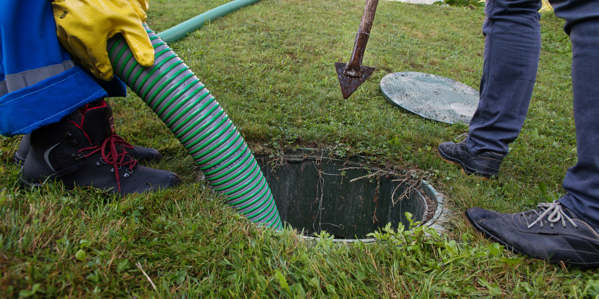 A septic tank is being serviced. A worker in yellow gloves handles a green hose near an open tank on a grassy lawn.