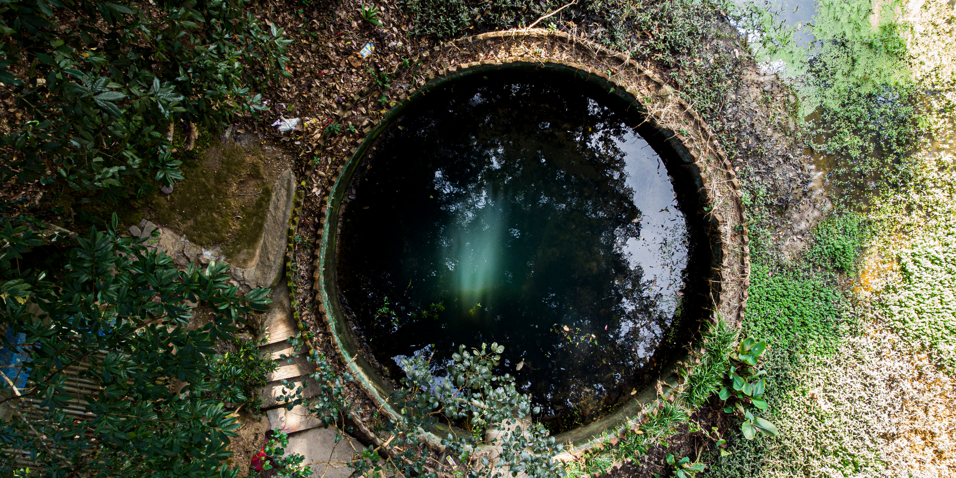 Overhead view of a circular well surrounded by greenery. Water reflects sky.